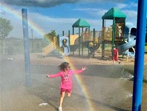 A young girl joyfully splashes in the water, surrounded by a vibrant rainbow arching overhead.