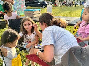 A woman face painting kids, taking in the fun & excitement of a community event.